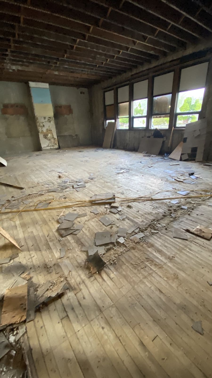 School interior mid-abatement — lifted floor tile and fragments across the wood subfloor, window band along the wall